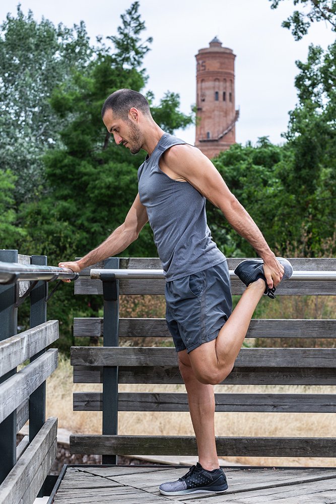 A person with light blonde hair is drinking from a shaker bottle in a gym. The gym has bright red walls and motivational posters on them. The floor features green turf, and there are various pieces of gym equipment in the background.
